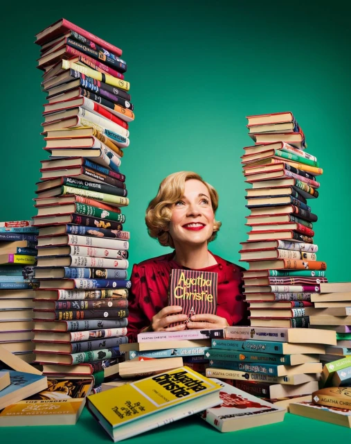 A woman with blond hair smiles, holding an Agatha Christie book, surrounded by large stacks of hardcover books against a green backdrop.
