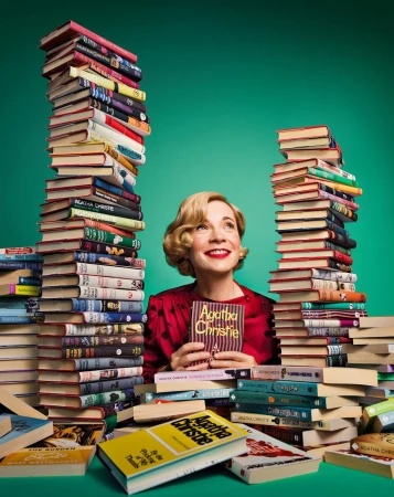 A woman with blond hair smiles, holding an Agatha Christie book, surrounded by large stacks of hardcover books against a green backdrop.