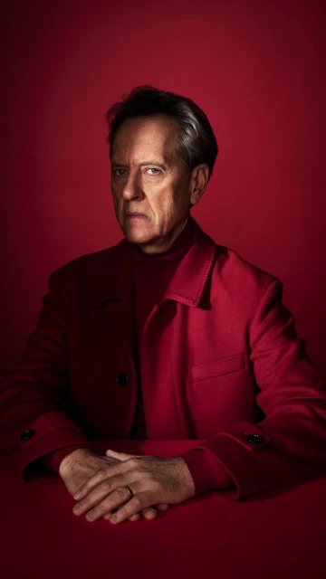 Portrait of a mature adult man with grey hair wearing a red turtleneck sweater and jacket, seated at a red table against red backdrop.
