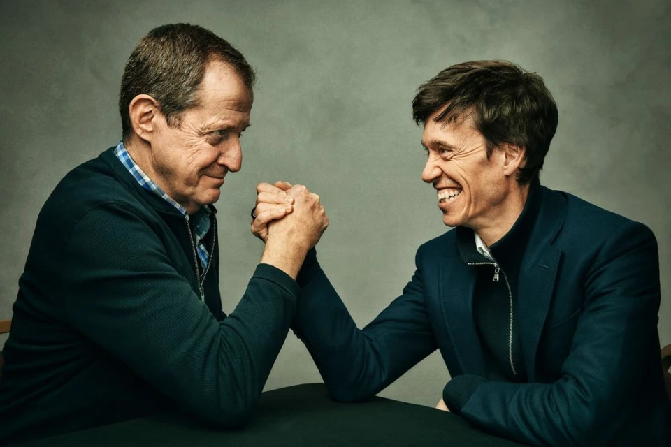Two adult men are engaged in arm wrestling at a table. One smiles, dressed in a blue jacket, while the other wears a blue sweater.