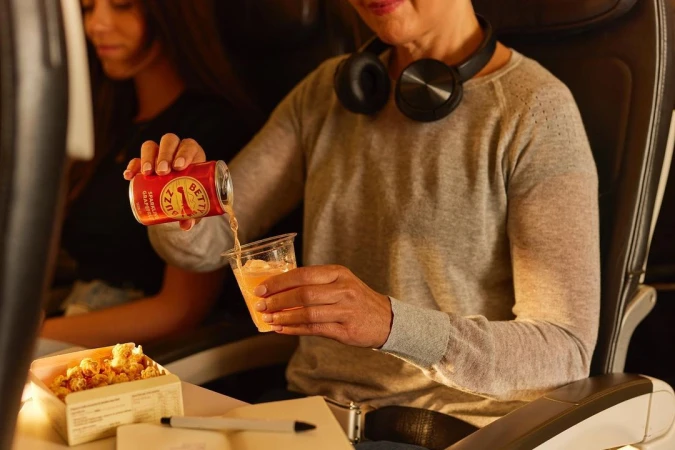 Woman pours sparkling grapefruit beverage from can into plastic cup on plane; popcorn, headphones, and in-flight details visible.