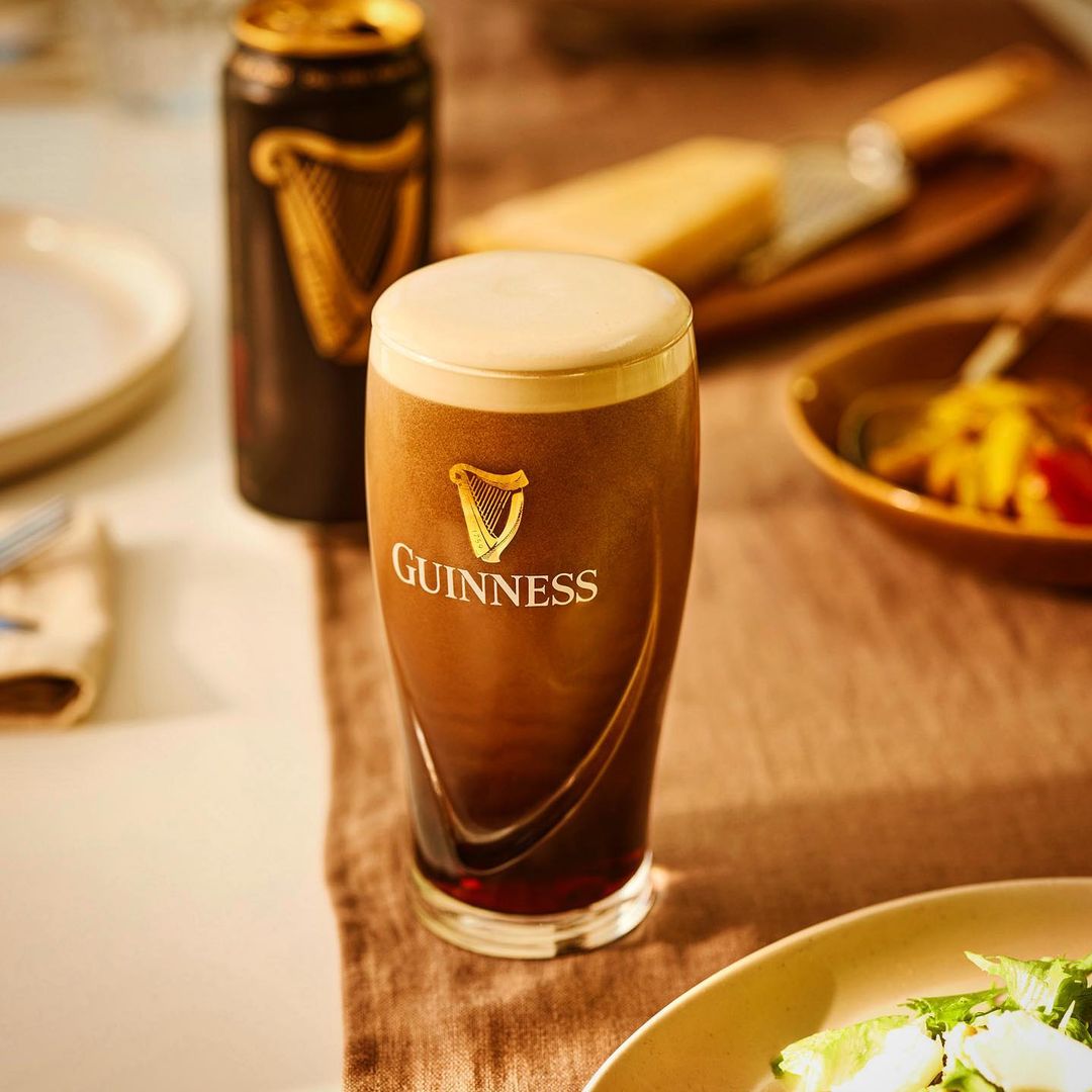 Guinness stout in glass next to a can, served with salad and cheese. The scene is staged on a light-colored table indoors.