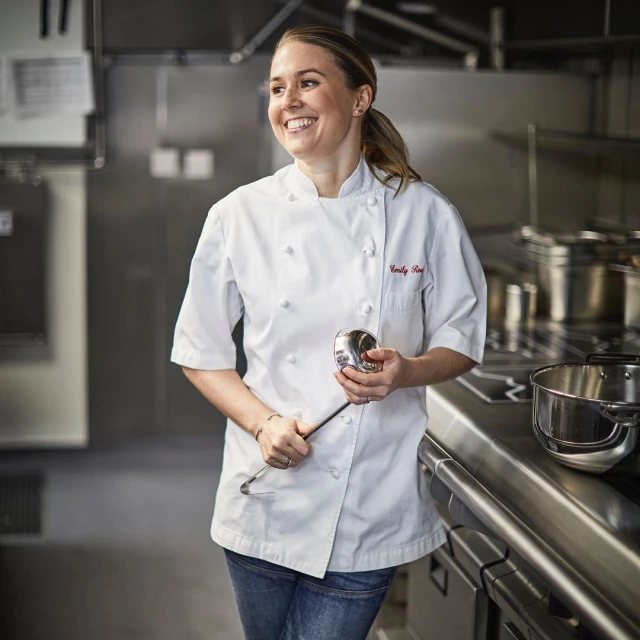 Smiling chef in white uniform in a commercial kitchen, holding a ladle, standing next to a stainless steel counter and cooking equipment.