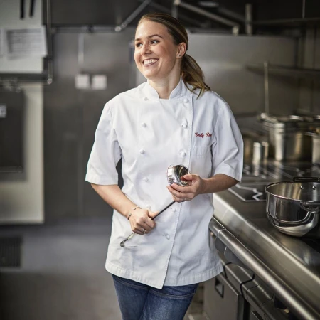 Smiling chef in white uniform in a commercial kitchen, holding a ladle, standing next to a stainless steel counter and cooking equipment.