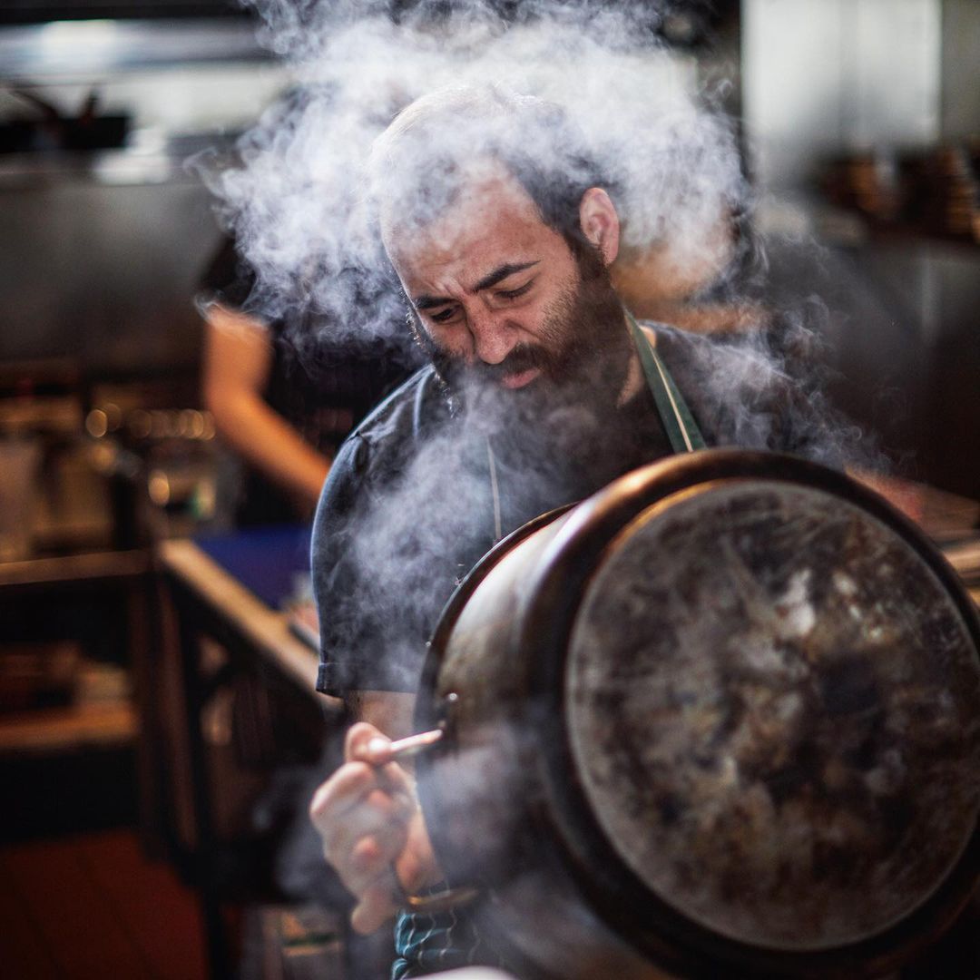 Chef with a beard cooking in a commercial kitchen with steam rising from a large pot, captured in a medium shot.