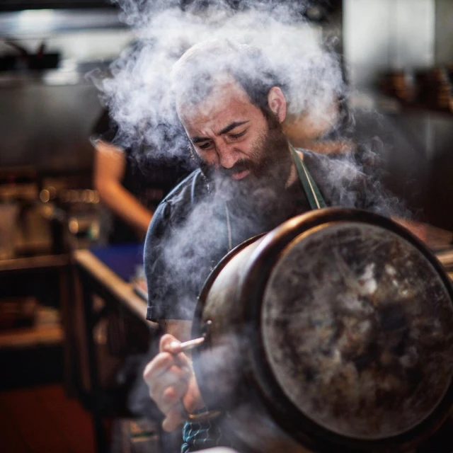Chef with a beard cooking in a commercial kitchen with steam rising from a large pot, captured in a medium shot.