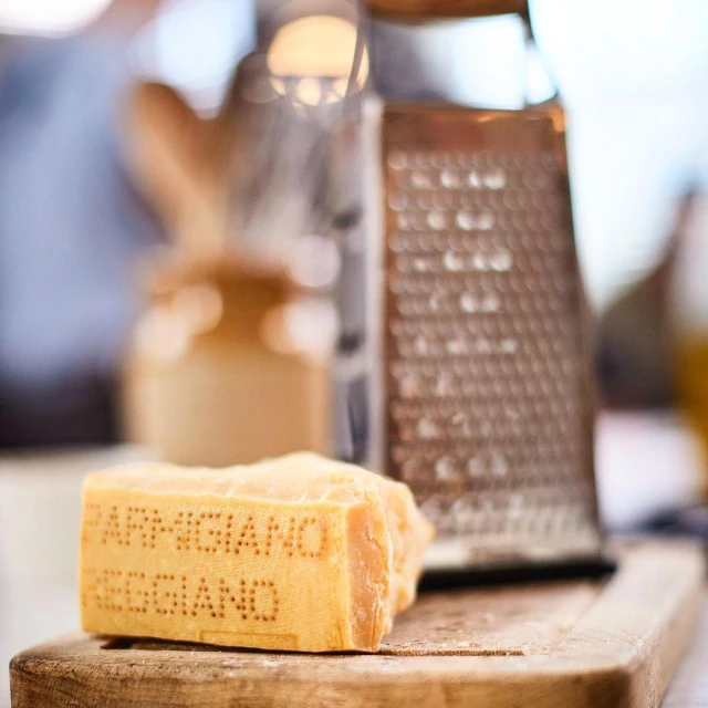 Close-up of block of Parmigiano Reggiano cheese with text on a wooden board next to a metal cheese grater in soft, diffused light.