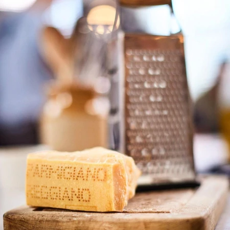 Close-up of block of Parmigiano Reggiano cheese with text on a wooden board next to a metal cheese grater in soft, diffused light.