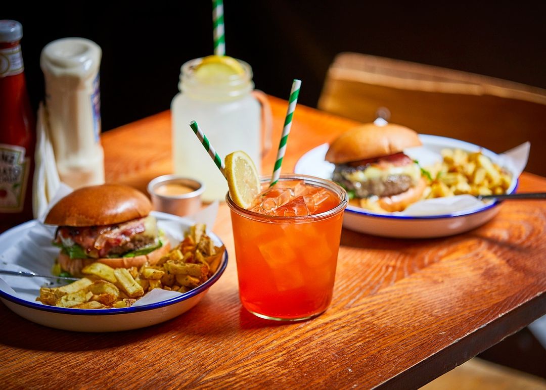 Two burgers and french fries sit on plates next to drinks with straws on a wooden table with ketchup and mayonnaise in the background.