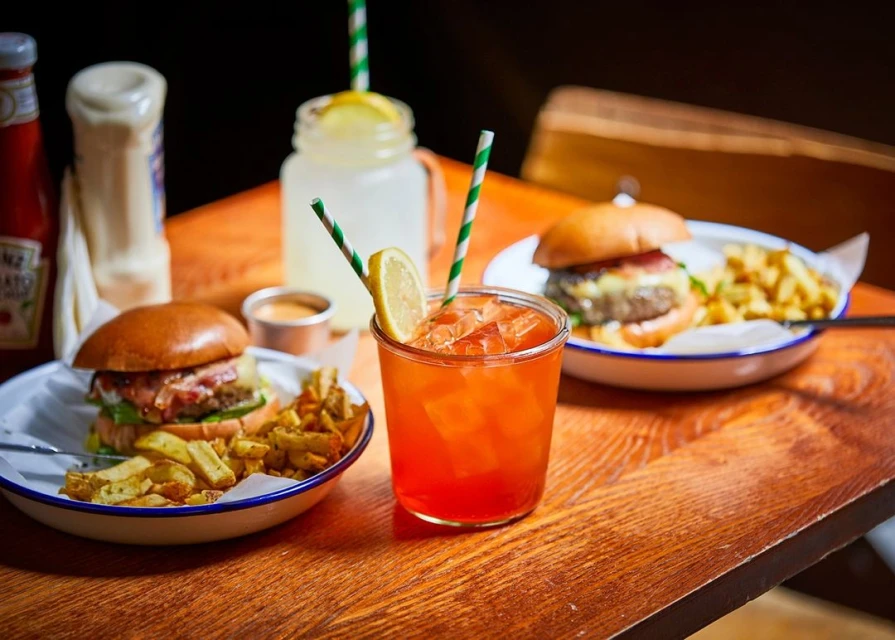 Two burgers and french fries sit on plates next to drinks with straws on a wooden table with ketchup and mayonnaise in the background.