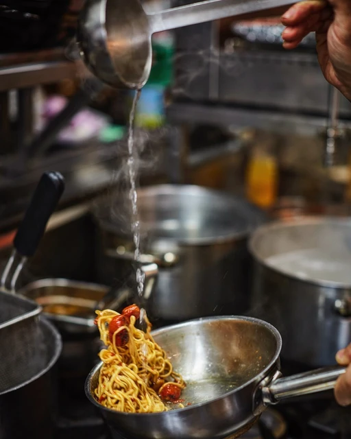 Spaghetti with tomatoes cooking with a ladle pouring water into a metal pan on a stovetop in a commercial kitchen setting.