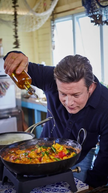 Chef Jamie Oliver pours oil on vegetables in a wok on a stove in a bright indoor kitchen setting, preparing food for cooking.