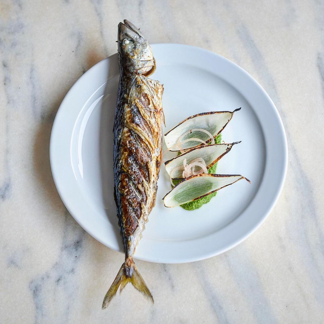 Overhead view of grilled whole fish on a white plate, served with onion slices and green sauce, against a marble surface.