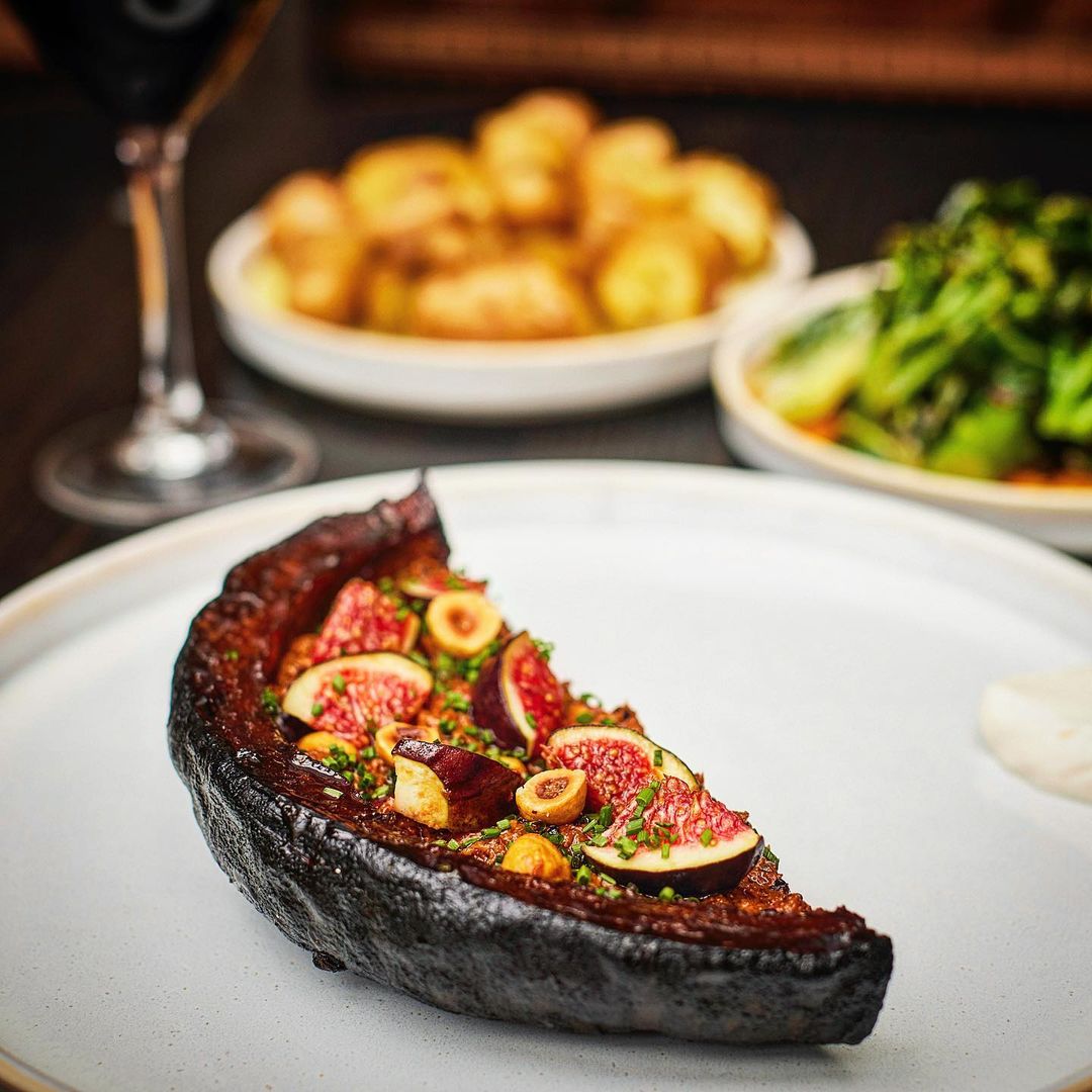 Close-up of a gourmet fig appetizer on a white plate, accompanied by roasted potatoes, green vegetables, and a glass of red wine.