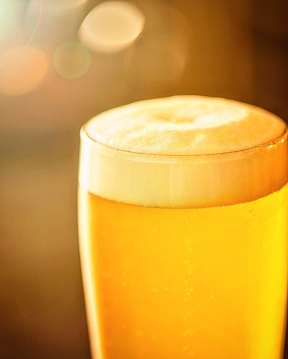 A close-up shot of a tall glass of golden beer with a thick white head, illuminated by a warm, diffused light, creating a bokeh effect.