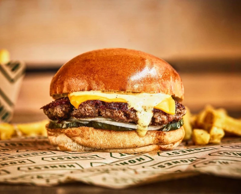 Studio shot of a cheeseburger with dripping cheese on a sesame bun, sitting on a branded paper, with french fries in the background.