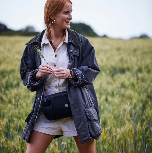 A redhead woman stands in a wheat field, wearing an olive green jacket and carrying wheat stalk in a medium shot, smiling gently.