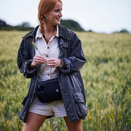 A redhead woman stands in a wheat field, wearing an olive green jacket and carrying wheat stalk in a medium shot, smiling gently.