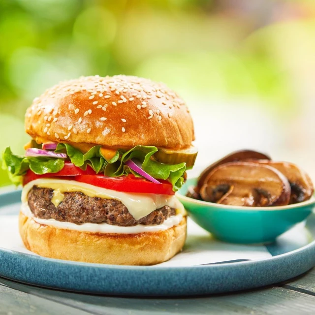 Close-up of a juicy burger with tomato, cheese, lettuce, and pickles on a sesame bun, next to mushrooms on a bright, sunny day.
