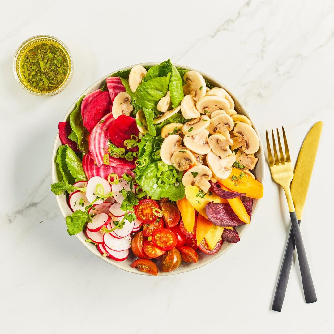 Overhead shot of a colorful salad in a white bowl with golden fork and knife on a marble surface, beside a bowl of pesto sauce.