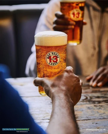 Two hands hold up glasses of amber-colored beer with white foam, showcasing the Hop House 13 Lager logo, set on a rustic wooden table.