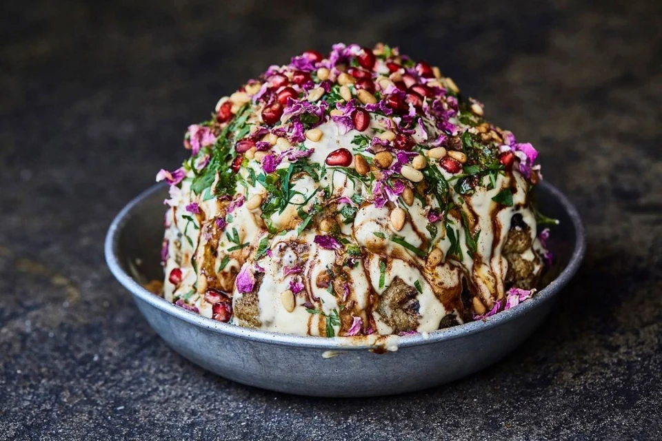 Close-up of a whole roasted cauliflower topped with a creamy sauce, pomegranate seeds, parsley, pine nuts, and edible flowers in a metal bowl.