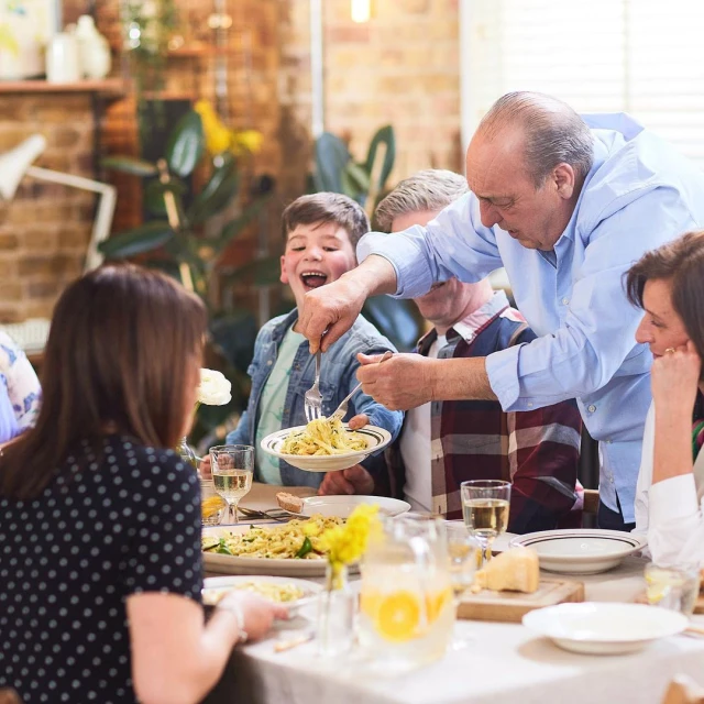 Family gathered around a dining table enjoying a meal with pasta, drinks, and bread in a warmly lit indoor setting, sharing food.