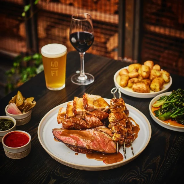 Overhead shot of a gourmet meal with grilled meat, vegetables, beer and wine on a dark table, against a textured background.