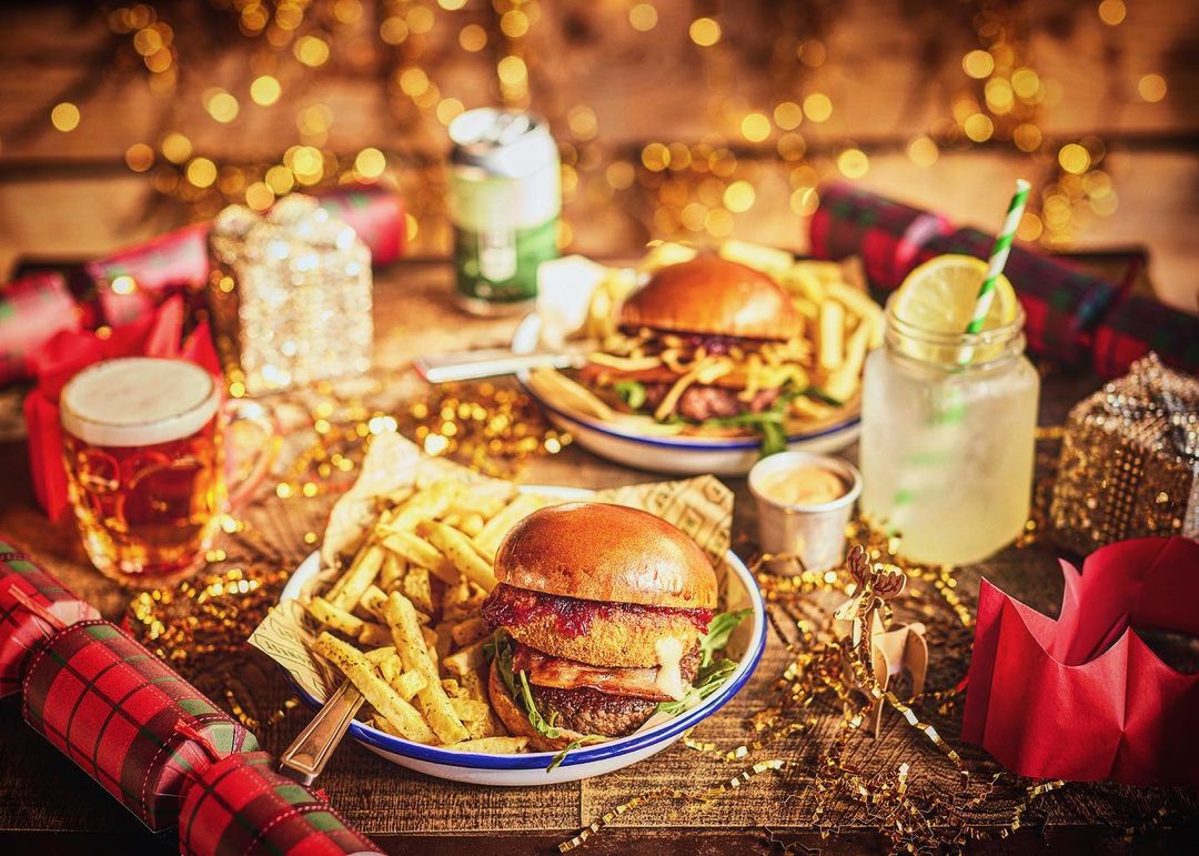 Festive food scene featuring burgers, fries, and drinks arranged on a wooden table with Christmas decorations and bokeh lights.