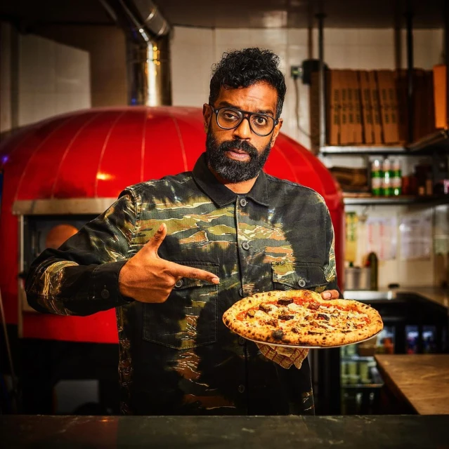 Adult male holding a pizza in front of a red pizza oven. He's wearing glasses and a patterned shirt, indoors, looking at the camera.