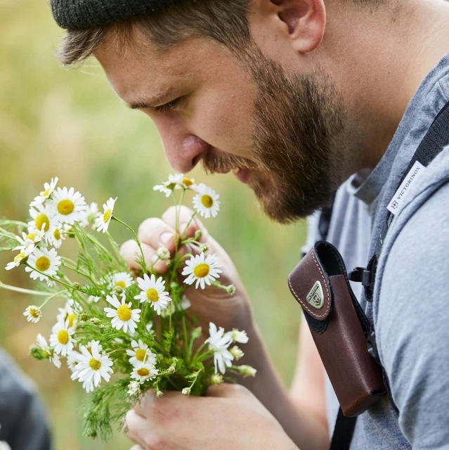 Man smelling bunch of daisies outdoors, wearing casual grey t-shirt and black beanie with leather pouch on his shoulder.