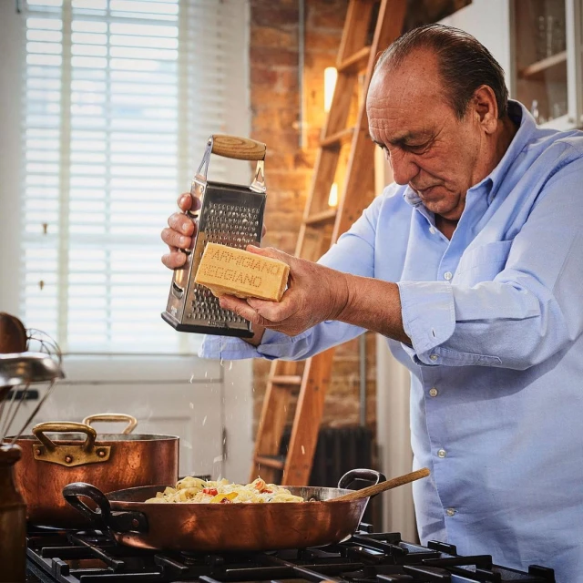 Adult male grating Parmigiano Reggiano cheese into a copper pan with food in an indoor kitchen setting with natural window light.