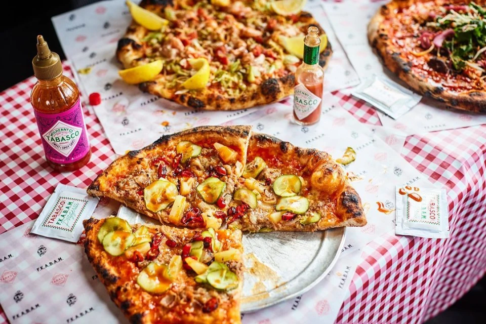 Three pizzas, one sliced, with Tabasco bottles and packets on a red gingham tablecloth, presented in a food and beverage setting.