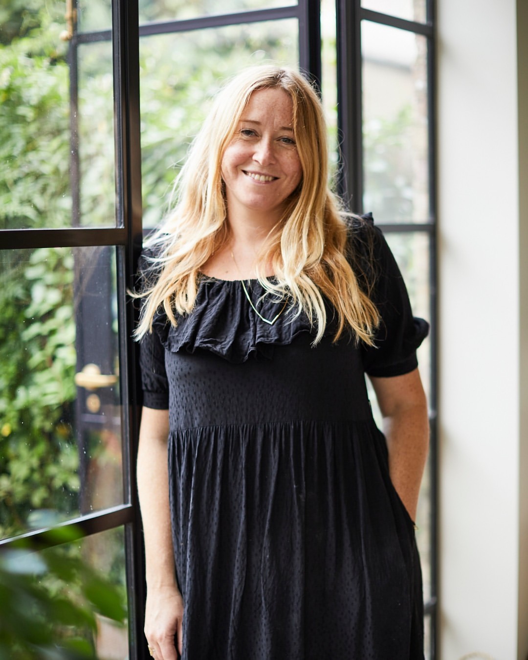 Blond woman in a black dress poses indoors near a dark-framed window with greenery, creating a portrait-style image with natural lighting.