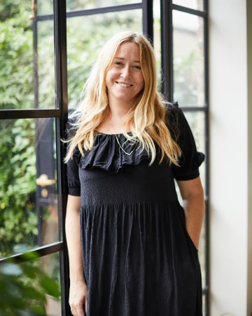 Blond woman in a black dress poses indoors near a dark-framed window with greenery, creating a portrait-style image with natural lighting.
