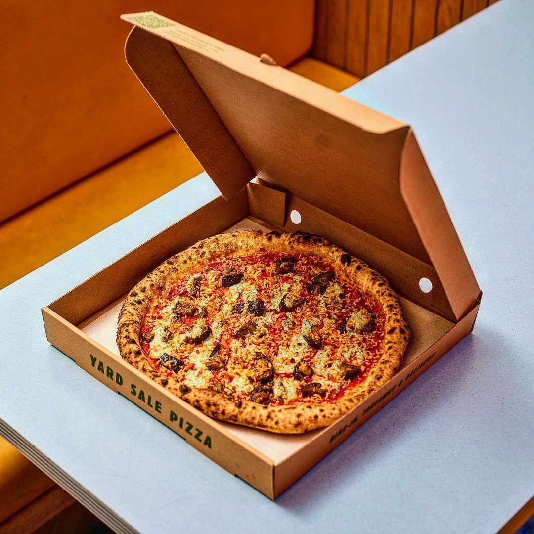Overhead studio shot of a delicious Yard Sale Pizza in its cardboard box, with visible cheese, toppings, and golden-brown crust.