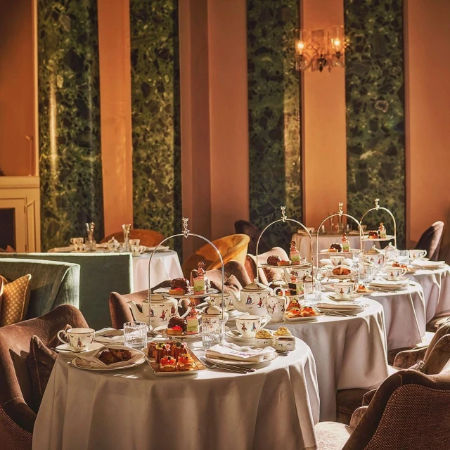 Elegant afternoon tea setting with tiered stands of pastries and teacups on white tablecloths, set against a sophisticated interior backdrop.