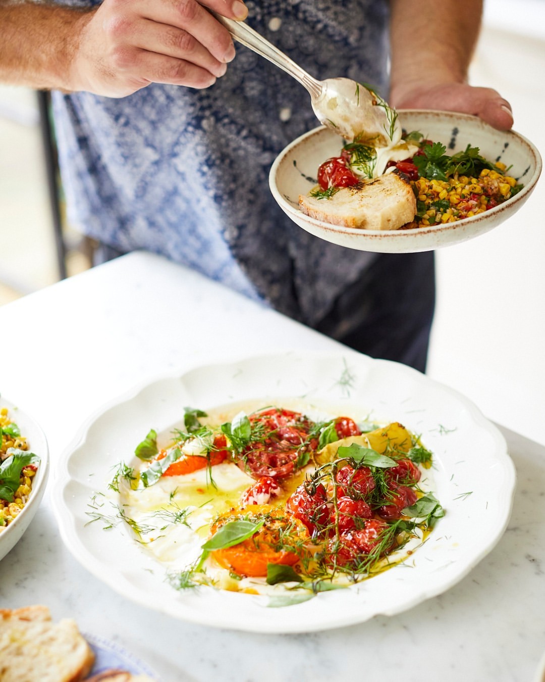 Overhead shot of an adult preparing and serving tomato and basil salad with a silver spoon on a white marble countertop indoors.