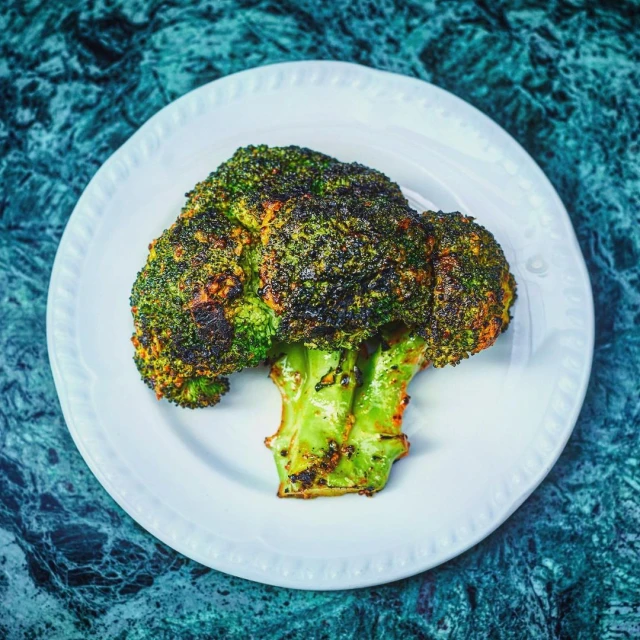 Overhead shot of charred green broccoli on a round white plate, centered against a mottled teal and blue surface.