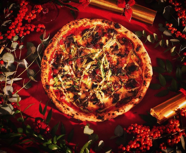 Overhead shot of a pizza on a red tablecloth, garnished with greenery and red berries, flanked by golden party crackers and wine glass.
