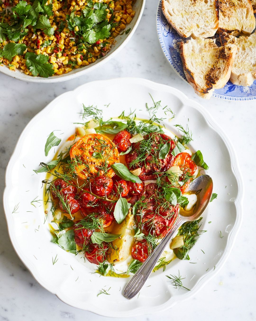 Overhead shot of a white plate with heirloom tomatoes, fresh herbs, and a silver spoon, with corn salad and toast in the background.