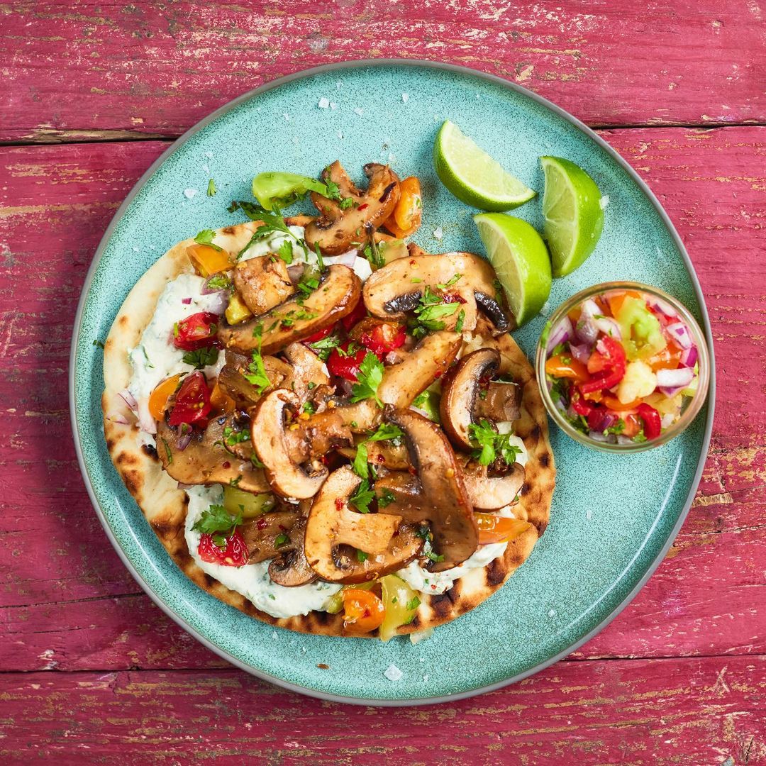 Overhead shot of a plate featuring a flatbread topped with sauteed mushrooms, tomatoes, and herbs, accompanied by lime wedges and salsa.