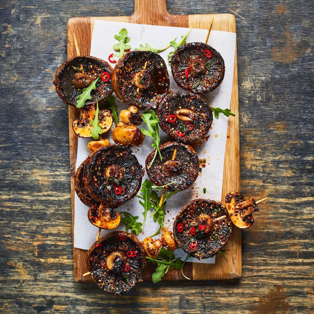 Overhead shot of grilled portobello mushroom skewers with greens and red pepper flakes, displayed on a wooden board for food presentation.