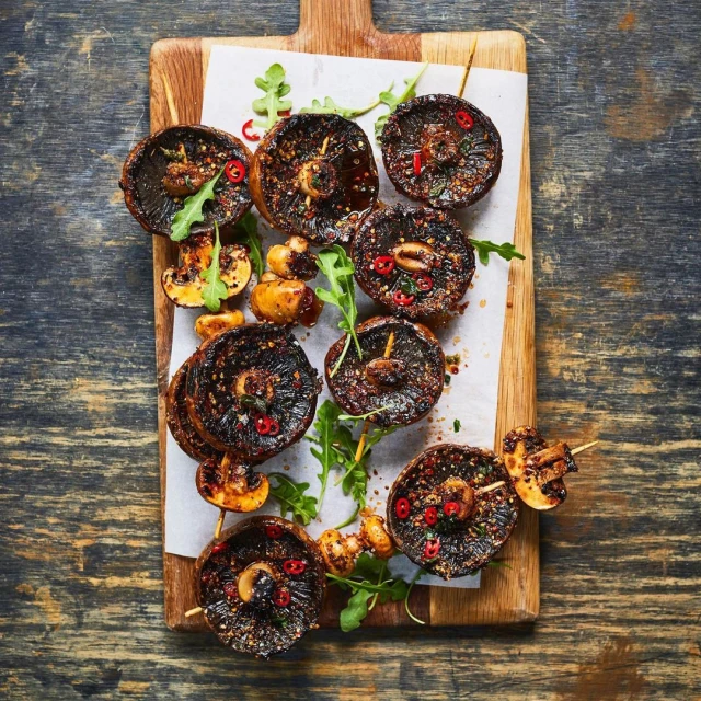 Overhead shot of grilled portobello mushroom skewers with greens and red pepper flakes, displayed on a wooden board for food presentation.