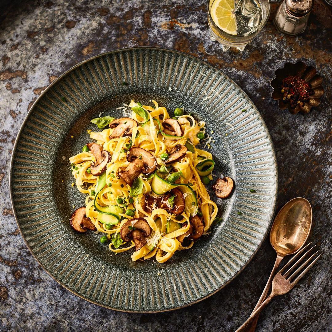 Overhead shot showcases a plate of fettuccine primavera with mushrooms, zucchini, peas, herbs, and grated cheese, paired with a glass of water.