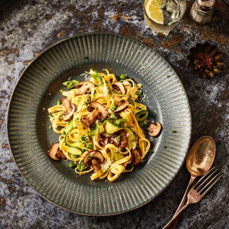 Overhead shot showcases a plate of fettuccine primavera with mushrooms, zucchini, peas, herbs, and grated cheese, paired with a glass of water.