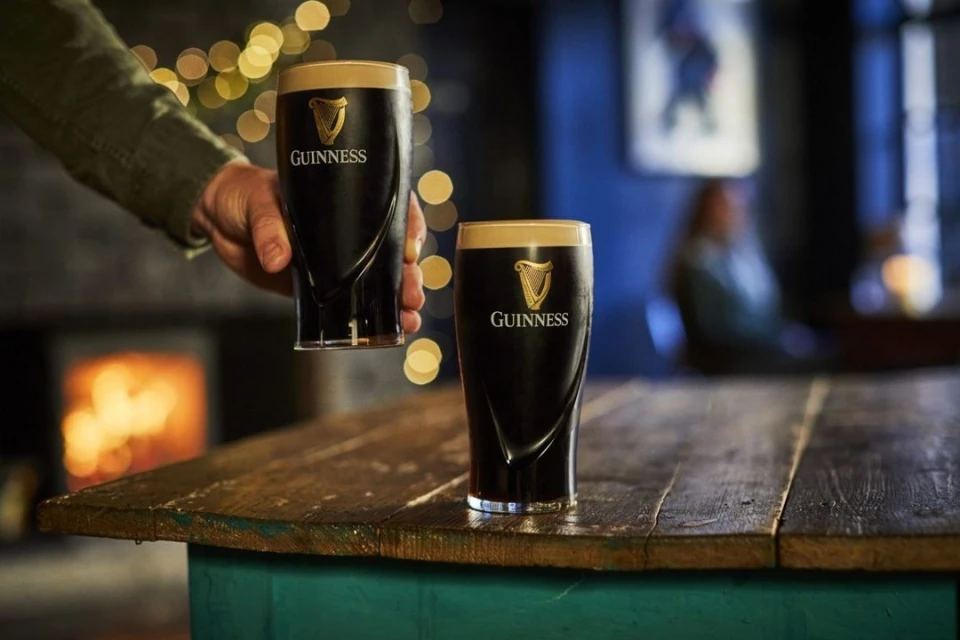 Two glasses of Guinness stout on a wooden table, one held by a person, against a blurred background with a fireplace.