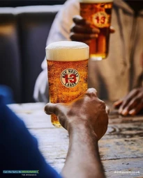 Close-up of a person holding a pint glass of Hop House 13 lager with a thick white head in a pub.