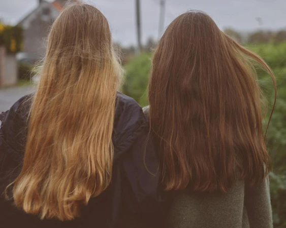 Two young women seen from behind with long flowing hair walking outdoors against a green background by photographer Sophie Van Der Perre.