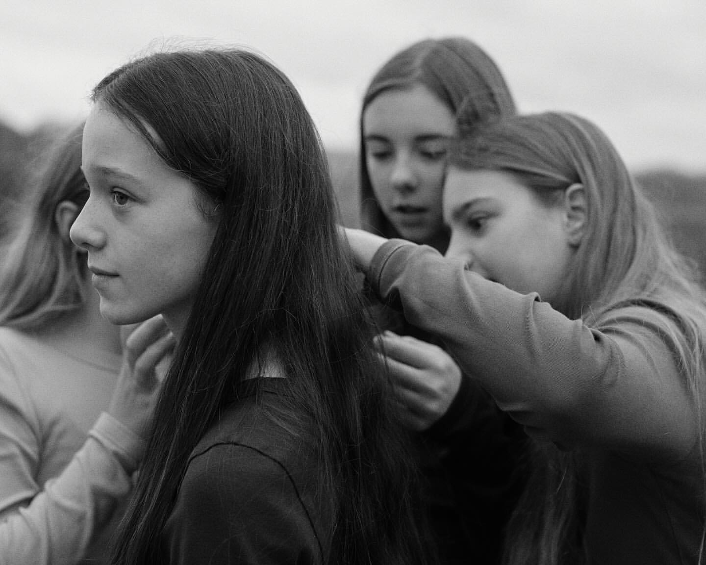 Three young women are outdoors in a black and white portrait. One is getting her hair styled by the other two, who are standing behind her.